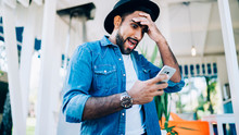 Stunned Hipster Guy Shocked From Bad Memory And Forgetting Stuff While Reading Received Email On Smartphone Gadget, Amazed Man Emotionally Holding Head And Checking Account Balance Of Sim Card