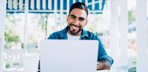 Portrait of cheerful Turkish software developer sitting at desktop with laptop device and smiling at camera while waiting for technology updating, happy man in casual wear working remotely