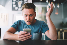 Amazed Hipster Guy With Cellphone Gadget In Hands Feeling Shocked From Notification On Application Sitting At Cafe Interior With Wifi Internet, Wonder Caucasian Male Blogger Reading Text Comment