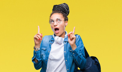 Wall Mural - Young braided hair african american student girl wearing backpack over isolated background amazed and surprised looking up and pointing with fingers and raised arms.