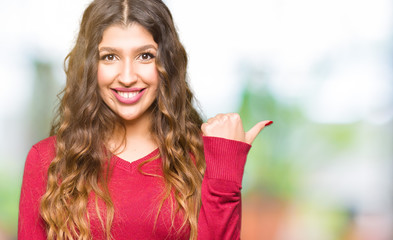 Young beautiful woman wearing red sweater smiling with happy face looking and pointing to the side with thumb up.