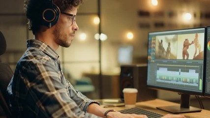 Wall Mural - evening in creative office: professional videographer / audio engineer wearing headphones works on d