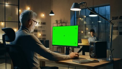 Sticker - Creative Middle Aged Designer Sitting at His Desk Uses Desktop Computer with Green Mock-up Screen. In the Evening Two Creative Employees Working on Computers in the Office with Cityscape Window View