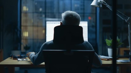Back view Shot of the Businessman Sitting at His Desk Using Desktop Computer. Stylish Office Studio with Dimmed Light and Big Cityscape Window View