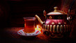 Hot Turkish glass of red tea with artistic teapot. Old Arabic Teapot with smoke over dark background. Arabic objects over Warm fabric. Middle east elements. Ramadan Background. Arabian Nights.