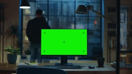 Desktop Computer with Mock-up Green Screen Standing on the Wooden Desk in the Modern Creative Office. In the Background Designer Drinks from a Cup Looks at the Night City out of the Window