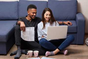 Image of young multicultural couples sitting on floor near blu sofa, using portabl compute for online working, two college students writting course work together, using lap top and wireless Internet.