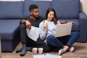 Interracial couple of young people sitting together in front laptop on floor near sofa, in their appartments, busy with composing financial report, using paper documents and portable computer,