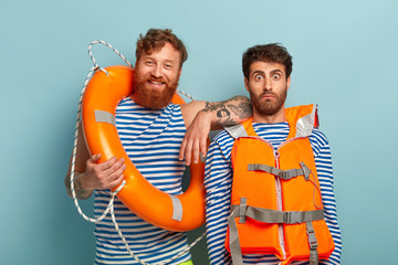 Happy red haired bearded male lifeguard and his colleague control safe swimming at sea, being glad and surprised, rescue people, use lifebuoy, wear striped jumper with blue stripes, life vest