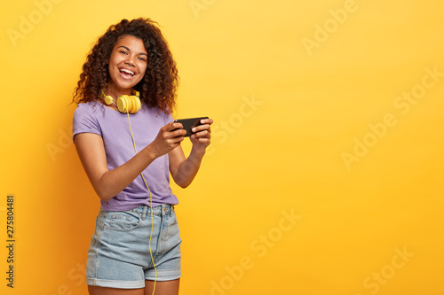 Half length shot of positive attractive female model with Afro haircut, feels good, uses smartphone device for entertainment and online chatting, surfes social network profile, uses free internet