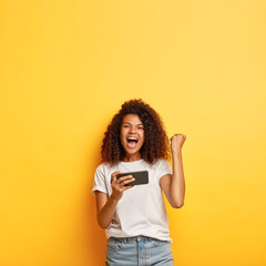 Excited cheerful dark skinned woman with bushy crisp hair, watches video online, clenches fist, celebrates winning online game chats in social networks wears fashionable outfit stands over yellow wall