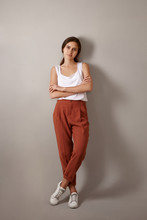 Vertical Full Length Studio Shot Of Trendy Looking Fashionable Young European Woman Dressed In White Top, Brown Trousers And Sneakers Posing Isolated, Crossing Arms Confidently, Enjoying Leisure Time