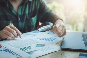 Business man using magnifying to review balance sheet annual with using laptop computer to calculating budget. audit and Check integrity before investment concept.