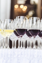 Several rows of red and white wine glasses on a table with white tablecloth skirt and blurred lights. Catering at wedding or occasion reception and party concept with copy space