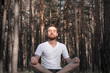 Young male person meditating in the forest. Modern man sits in the pine woods with closed eyes and enjoys the silence of nature