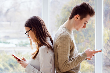 Couple of young students sitting next to one another and using mobile phones having no expression, concerned to their phones, have no interest in one another, problem of constant using modern gadgets