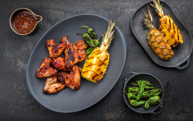 Wall Mural - Exotically barbecue chicken wings with hot chili sauce, jalapeno and pineapple as top view on a cast iron plate