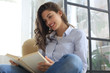 © ty - Smiling young female sitting in the armchair in the living room and reading book.