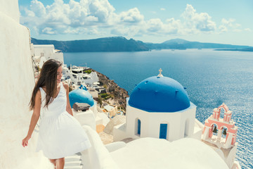 Fototapete - Santorini travel tourist woman on vacation in Oia walking on stairs. Asian girl in white dress visiting the famous white village with the mediterranean sea and blue domes. Europe summer destination.
