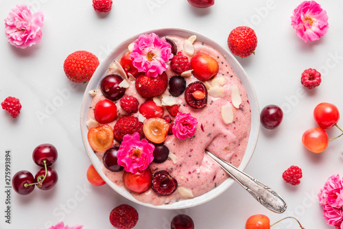 nice cream or smoothie bowl made of frozen bananas and berries with rose flowers, nuts and seeds with a spoon