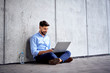 © baranq - Cheerful young businessman sitting outdoors in the city and working on laptop