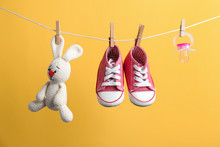 Small Shoes, Toy Bunny And Pacifier Hanging On Washing Line Against Color Background. Baby Accessories
