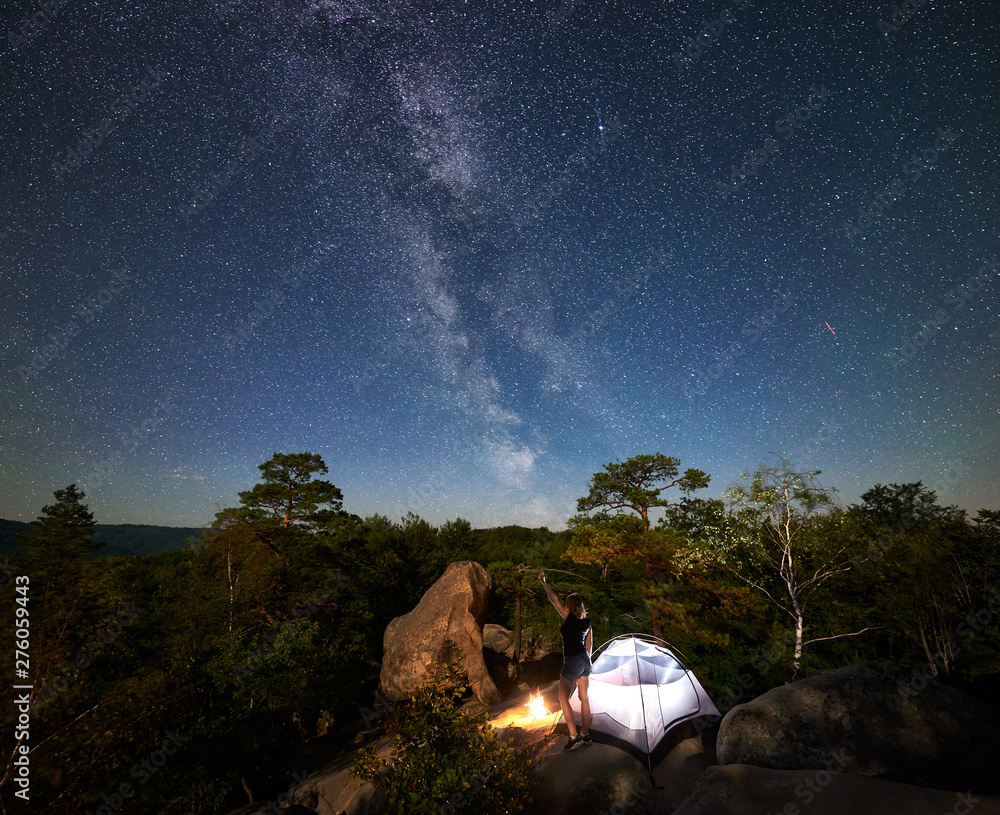 Rear view of woman hiker resting on rocky mountain top beside camp, campfire and tourist tent at summer night, pointing at sky full of stars and Milky way. On background starry sky, boulders and trees - obrazy, fototapety, plakaty 
