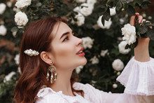 Outdoor Close Up Portrait Of Young Fashionable Happy Smiling Lady Wearing Trendy Vintage Style Earrings, White Pearl Barrette In The Long Hair, Posing In The Blooming Rose Garden