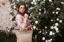 Outdoor Portrait Of Young Beautiful Fashionable Lady Wearing Pink Round Sunglasses, Dress, Holding Wicker Straw Basket Handbag. Model Posing In The Blooming Rose Garden. Copy, Empty Space