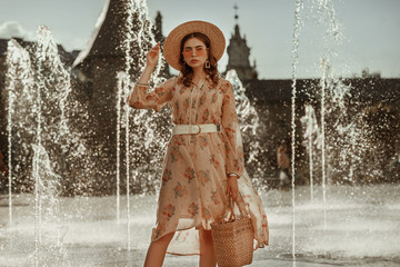 Outdoor portrait of young fashionable woman wearing straw hat, pink sunglasses, stylish dress with leather belt, holding straw wicker basket bag, posing in street of European city. Copy, empty space 