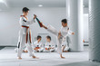 © Dusan Petkovic - Two young Caucasian boys in doboks having taekwondo training at gym. One boy kicking while other one holding kick target. In background their friend sitting with legs crossed and watching them.