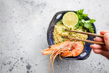 Wall Mural - A bowl of instant Chinese noodles with green onions, red hot chilli peppers, and shrimps on a gray stone background, top view