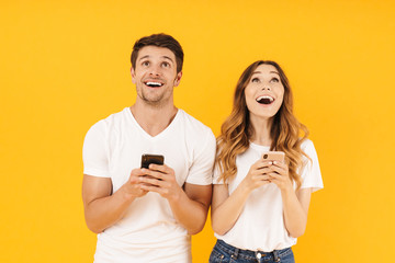 Wall Mural - Portrait of surprised couple man and woman in basic t-shirts looking upward together at copyspace while holding smartphones