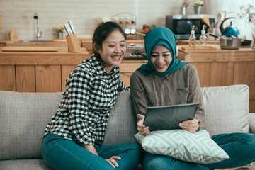 two beautiful young diverse women friends at home sitting on sofa using tablet computer and smiling. cheerful islam lady and roommate relax in couch on weekend day time watching movie on mobile pad.