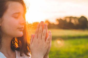 Wall Mural - Teenager Girl closed her eyes, praying in a field during beautiful sunset. Hands folded in prayer concept for faith, spirituality and religion. Peace, hope, dreams concept