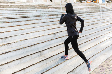 Wall Mural - Young muslim sports fitness woman dressed in hijab and dark clothes running outdoors at the street.