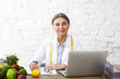 © shurkin_son - Picture of elderly mature female expert on diet and nutrition sitting at desk wearing white uniform and measure tape around her neck, using laptop, writing, making dieting plan for her client