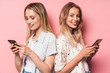 Cute cheerful beautiful blondes women friends posing isolated over pink wall background using mobile phones.