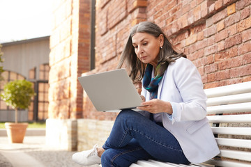 Wall Mural - Serious attractive businesswoman in white sneakers, jacket and blue jeans sitting on bench with portable electronic gadget on her lap, typing messages to business clients using 4g internet connection