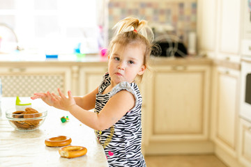 Little toddler kid having lunch in the warm sunny kitchen. Blonde girl with funny ponytail playing with two tasty bagels