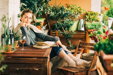 Wall Mural - Young beautiful woman relaxing on cozy balcony, reading a book, wearing warm knitted pullover, glass of wine on wooden table