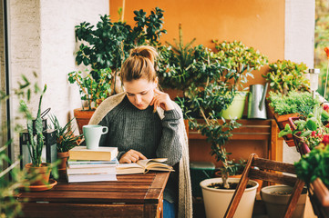 Wall Mural - Young beautiful woman relaxing on cozy balcony, reading a book, wearing warm knitted pullover, cup of tea or coffee on stack of books