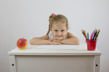 Portrait of a schoolgirl girl behind a white desk with colored pencils and an apple. Preschool education concept