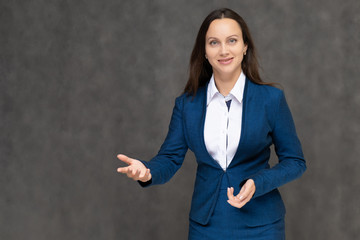 Portrait to the waist a young pretty brunette manager woman of 30 years in a business blue suit with beautiful dark hair. It is standing on a gray background, talking, showing hands, with emotions