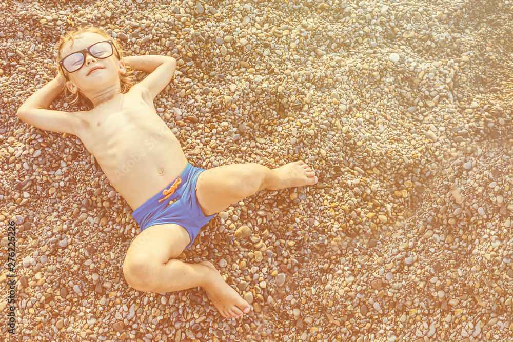 Cute funny little boy in big glasses sunbathing on the pebble beach ...