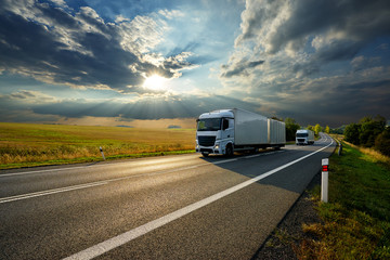 Wall Mural - two white trucks driving on the asphalt road in rural landscape at sunset with dramatic clouds