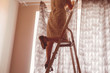 © Mihail - adult housewife hanging the curtains standing on the ladder at home