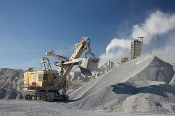 Wall Mural - Excavator and large heaps of gravel on the background of smoke from the pipe of the quarry crusher. Mining industry. Quarry and mining equipment.