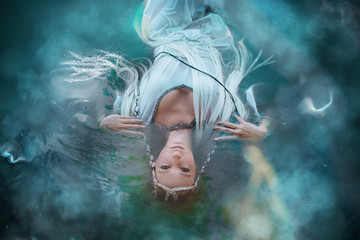 A girl elf in a white dress is floating in the water. Model in a medieval dress with pigtails floating in the lake