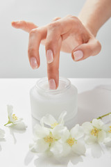 Wall Mural - cropped view of woman hand touching cream in jar near jasmine flowers on white surface